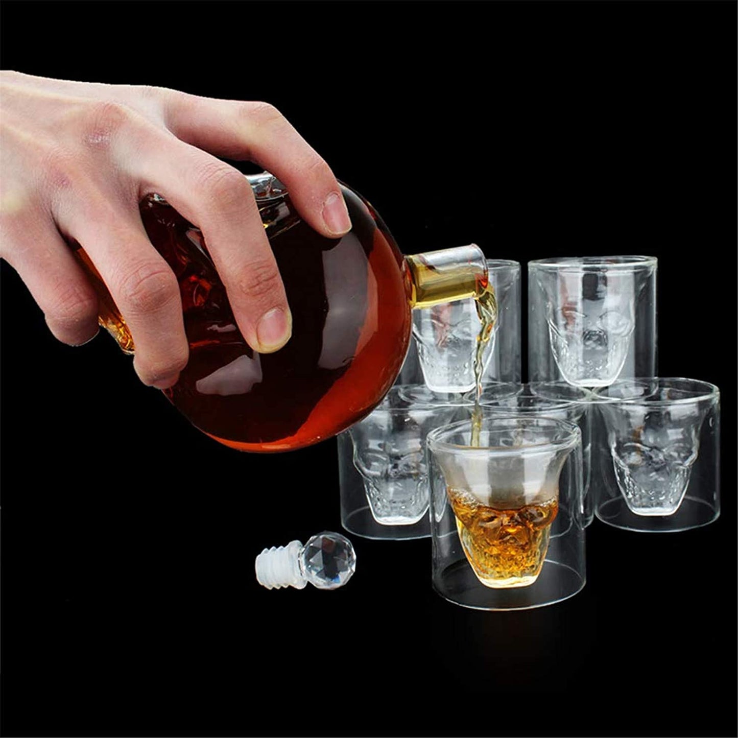 Hand pouring liquid from a bottle into skull-shaped glass cups on a black background