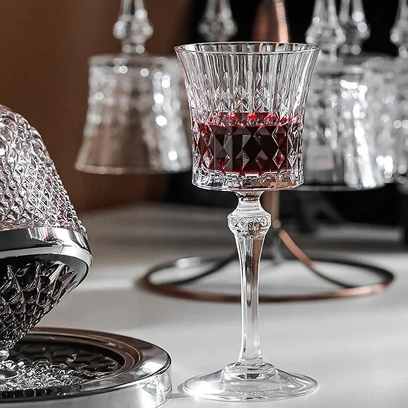 A clear cut-crystal wine glass with a stem and round base, partially filled with red wine, set on a tabletop with other matching crystal items blurred in the background.

