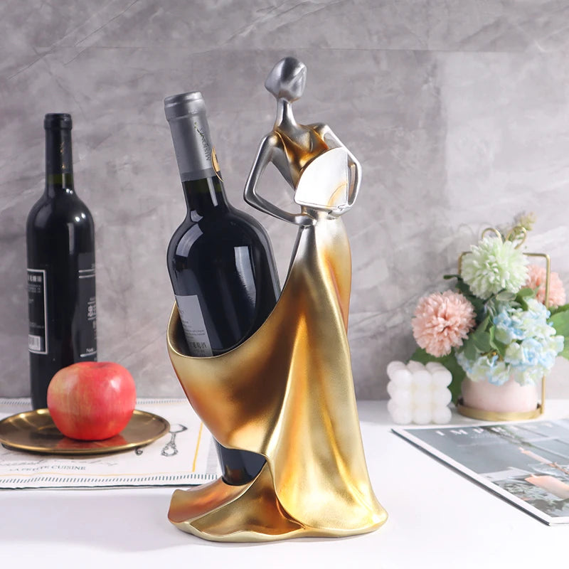 Decorative wine bottle holder shaped like a woman in a gold dress on a table with a bottle of wine and flowers.