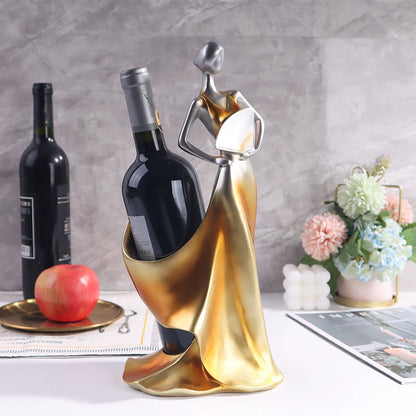 Decorative wine bottle holder shaped like a woman in a gold dress on a table with a bottle of wine and flowers.