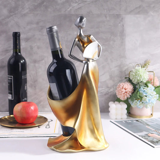 Decorative wine bottle holder shaped like a woman in a gold dress on a table with a bottle of wine and flowers.