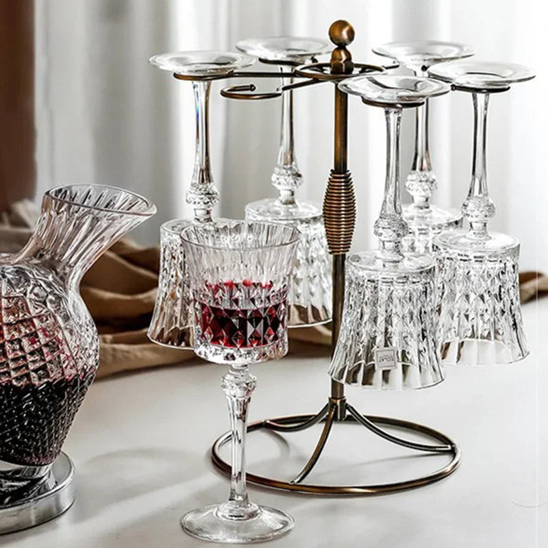 French-style 250ml crystal wine goblets with faceted stem and cut-crystal mounted on a finished metal frame,. A faceted glass decanter and a glass of red wine are partially visible in the foreground and background.


