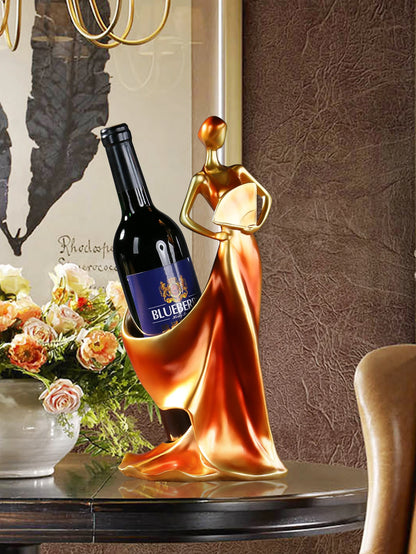 Decorative wine bottle holder shaped like a woman in a copper and gold dress, displayed on a table with flowers and a painting in the background.
