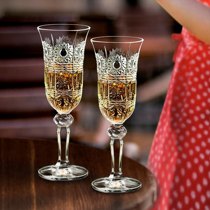 Two ornate crystal champagne flutes filled with sparkling champagne on a rustic wooden table, with a softly blurred figure in a red dress in the background, capturing an elegant celebration atmosphere and refined hosting moment.