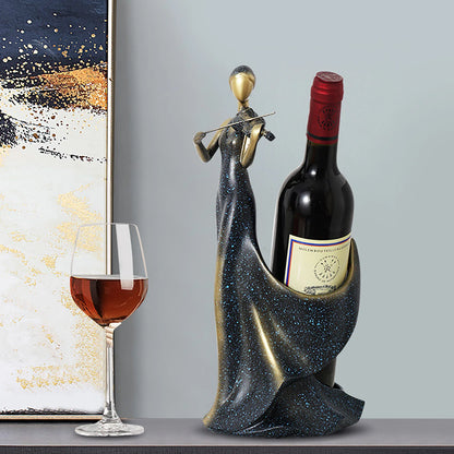 Decorative wine bottle holder shaped like a woman holding a violin with a bottle of wine and glass on a table.

