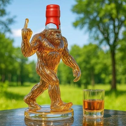 Glass bottle shaped like a Sasquatch with a red cap, next to a glass of amber liquid outdoors.